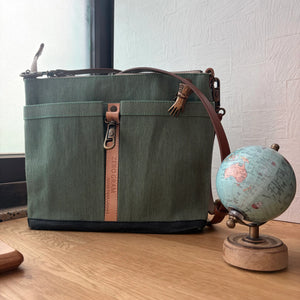 An olive green canvas shoulder bag with brown leather trim and a vertical 'ZERO GRAVITY' leather tab, displayed on a wooden table next to a small globe.