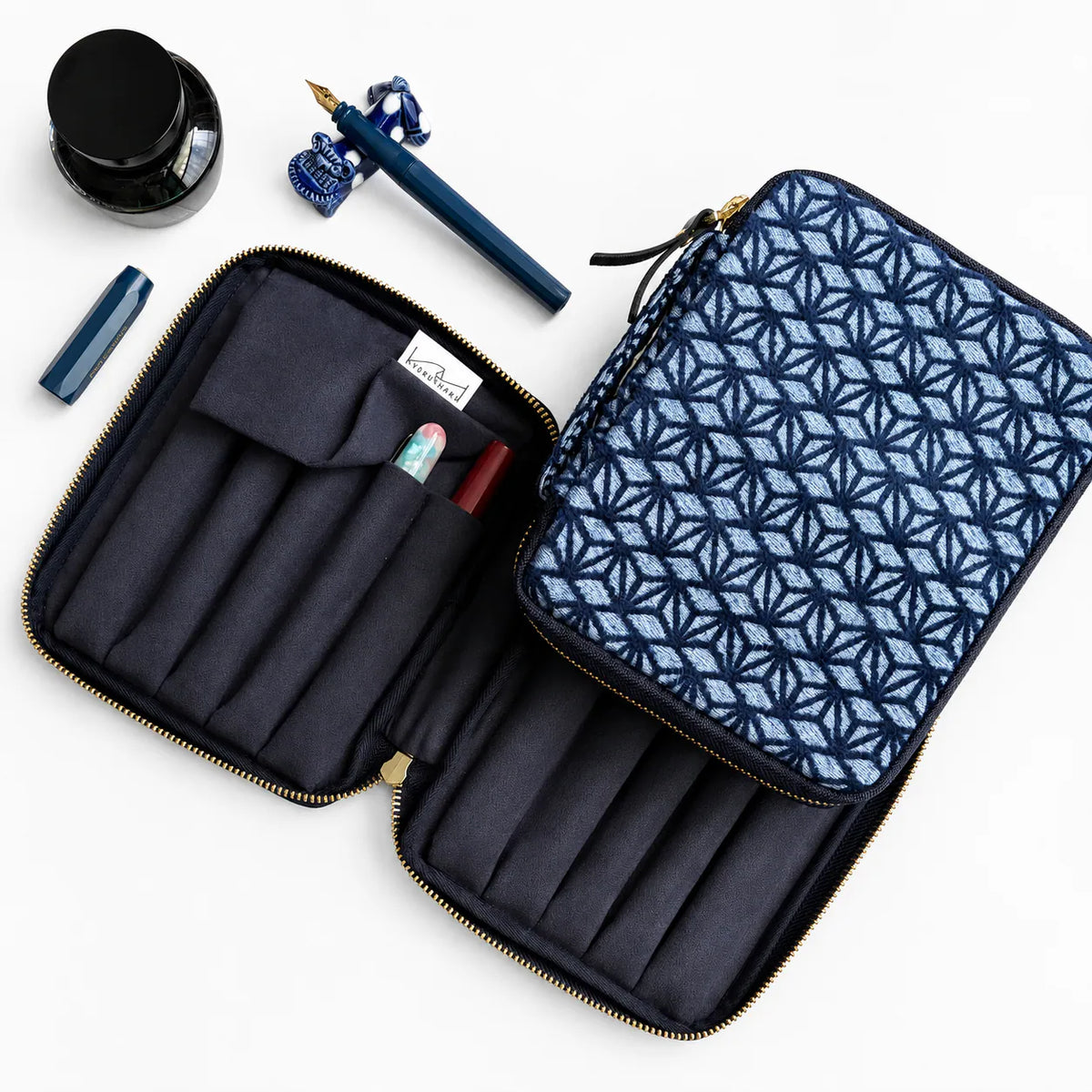 An open zippered travel organizer case with a navy and light blue geometric patterned exterior, dark navy interior with pen loops, shown on a white background with a fountain pen, ink bottle, and pen cap nearby.