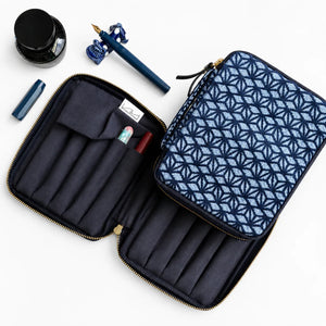 An open zippered travel organizer case with a navy and light blue geometric patterned exterior, dark navy interior with pen loops, shown on a white background with a fountain pen, ink bottle, and pen cap nearby.