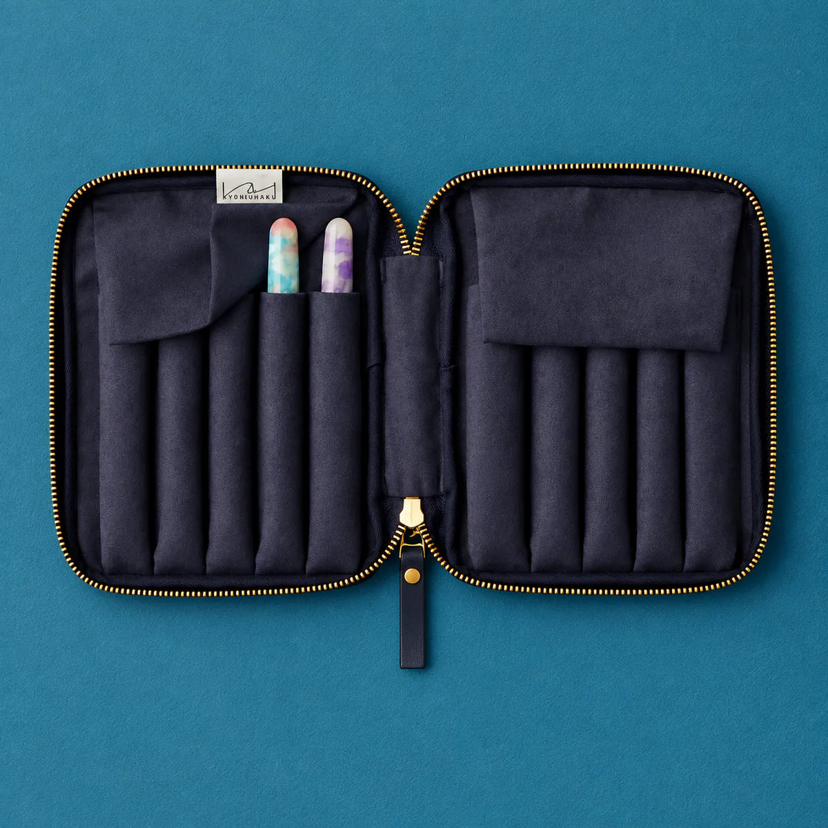 An open zippered jewelry organizer case in dark navy fabric with gold-tone zipper trim, laid flat on a teal background. The case has multiple vertical slots, a flap pocket, and two small gemstone or crystal pieces stored inside.