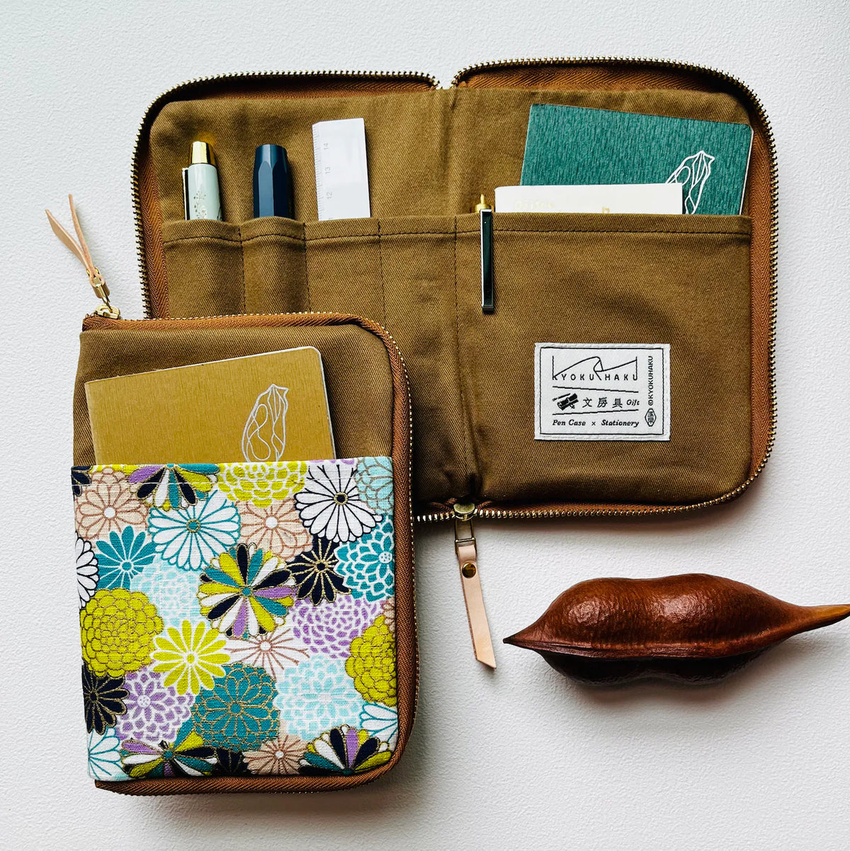 An open brown canvas zippered organizer pouch with multiple interior pockets, shown alongside a matching closed pouch featuring a colorful floral print. A small wooden leaf-shaped object sits to the right on a light neutral background.