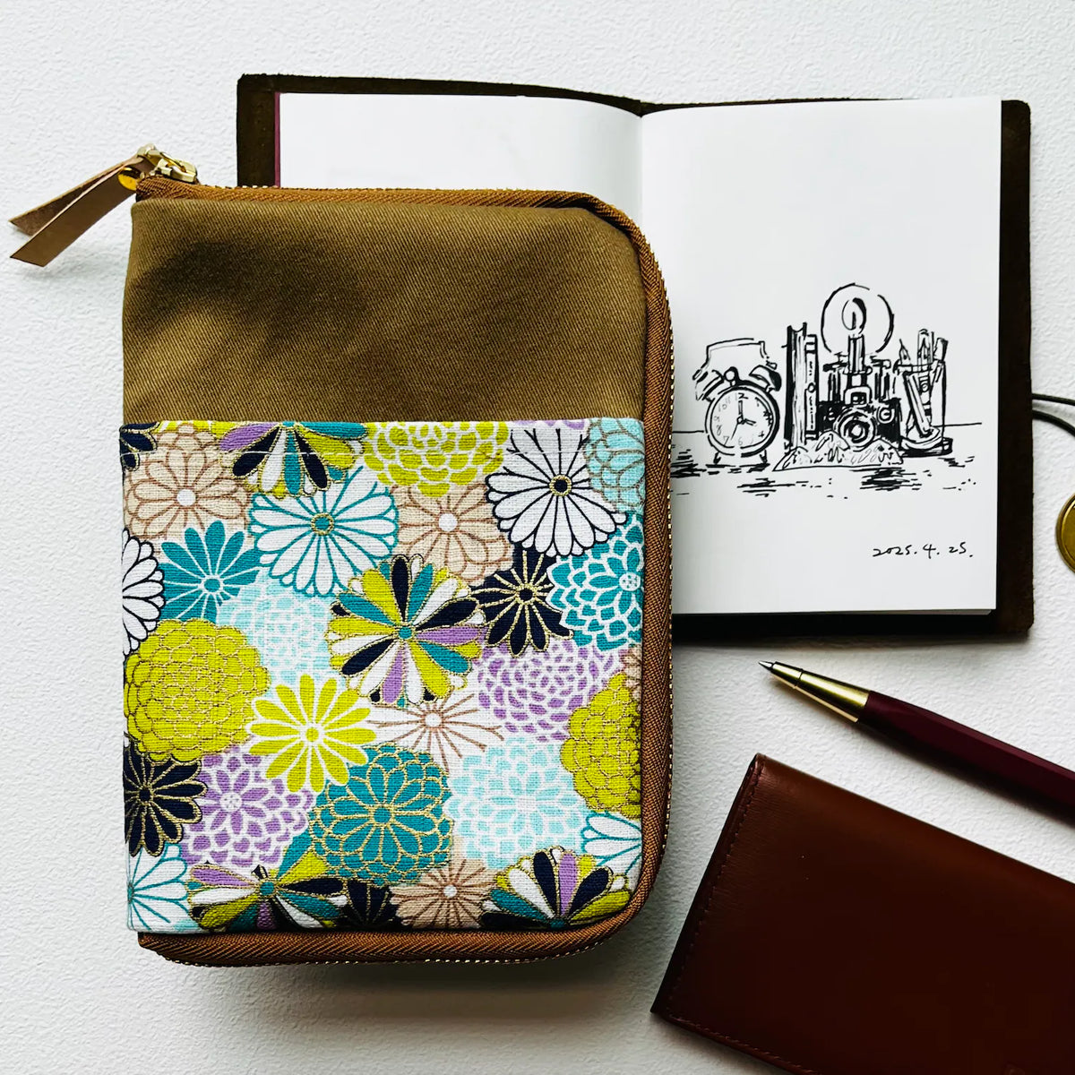 A zippered fabric organizer pouch with a brown top and bottom and a colorful floral patterned center panel, styled on a white surface next to an open notebook, a pen, and a brown leather wallet.