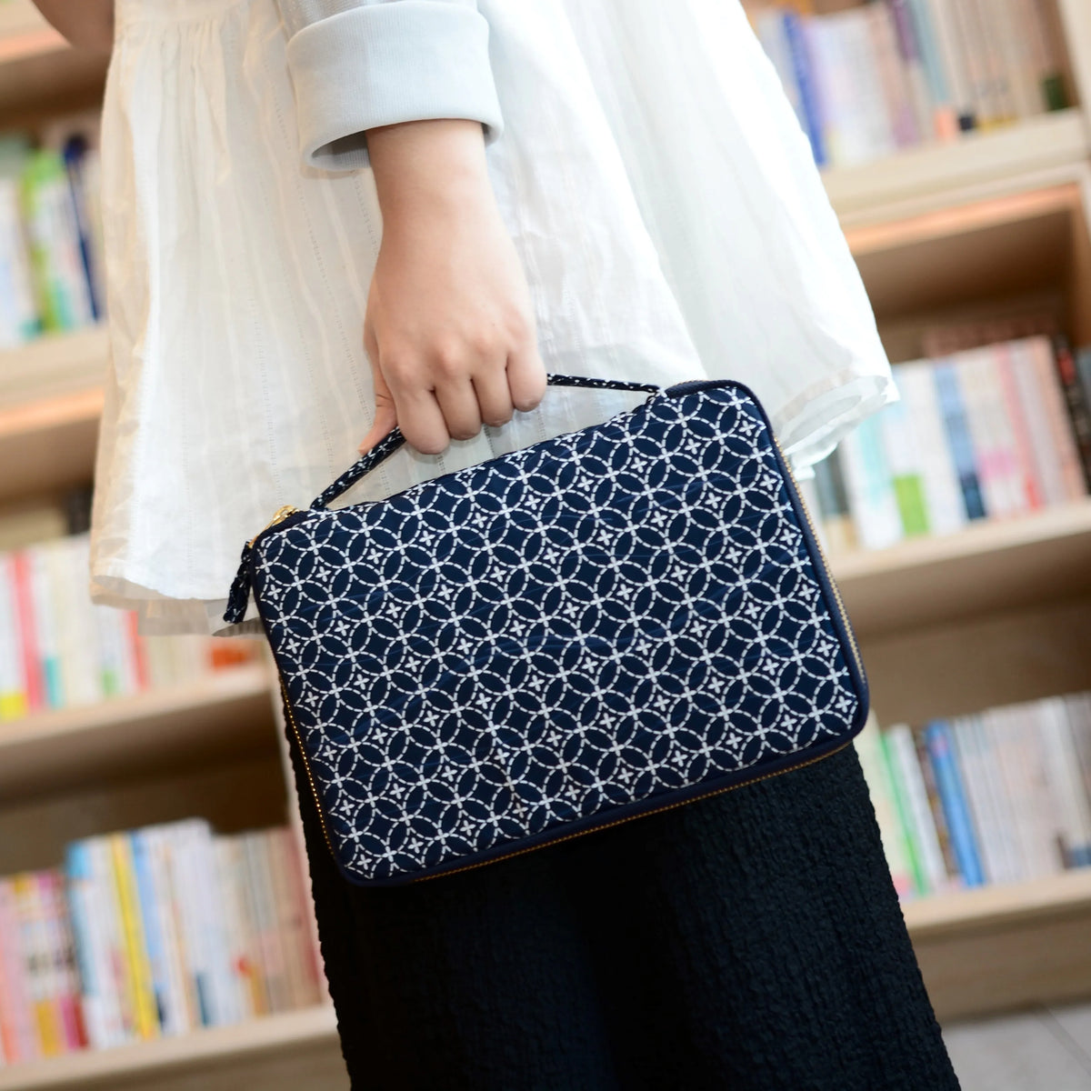 Person holding a navy blue patterned clutch in front of a bookshelf.