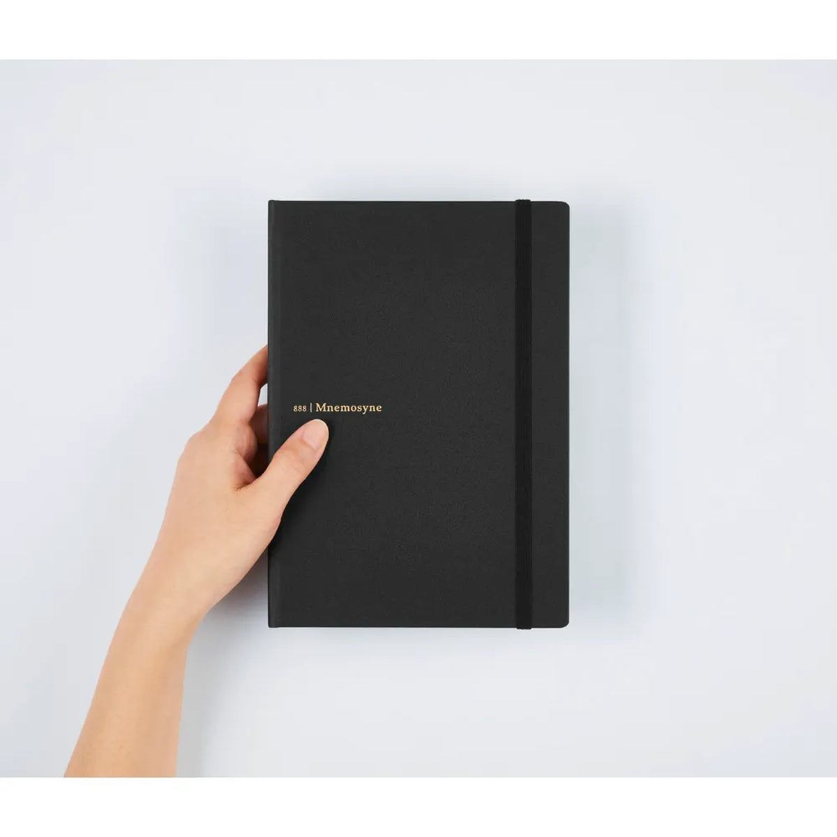 A hand holding a black notebook with a black elastic closure band and small gold foil text reading 'en | Macmoyne' on a light gray background.
