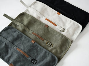 Four canvas pouches or sleeves arranged diagonally on a white background, shown in dark gray, olive green, off-white, and black. Each has a brown leather strap detail with a metal buckle.