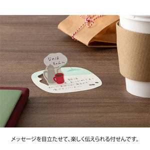 Sticker with a cat design and text on a wooden surface next to a coffee cup.