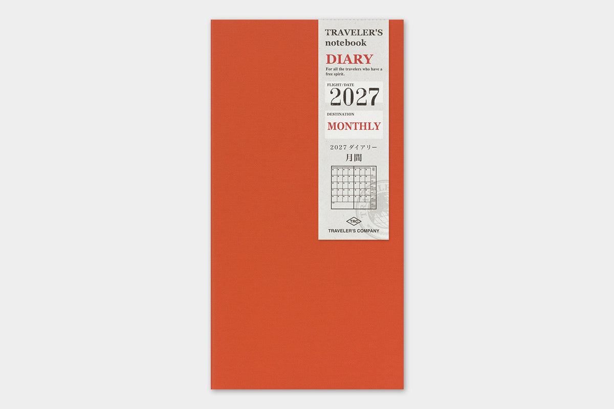 A vertical orange-red traveler's notebook with a white paper label on the front showing '2027 Monthly' and 'Traveller's Notebook' text, photographed against a light gray background.