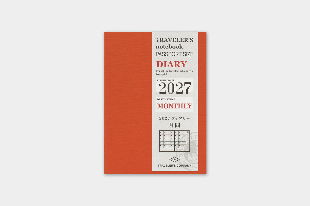 A vertical traveler's notebook with a coral orange cover and a white front panel showing '2027 Monthly' text and a small calendar graphic, photographed on a light gray background.