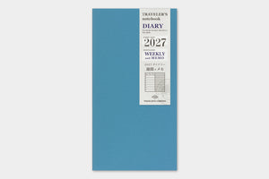 A blue traveler's notebook with a white paper cover label showing '2027' and weekly memo details, photographed against a light gray background.